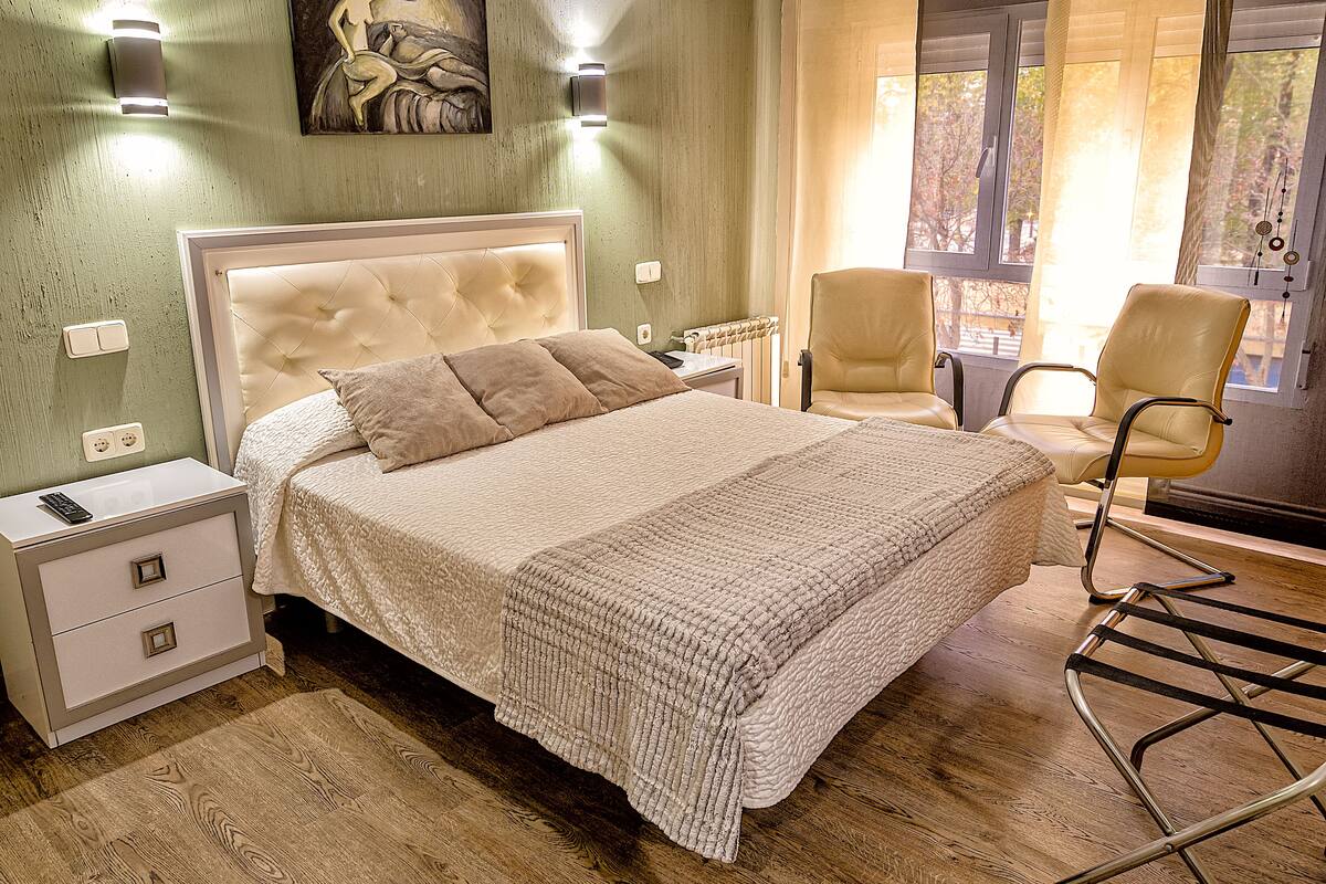 A bedroom with a bed, two chairs, a nightstand, and a painting on the wall.