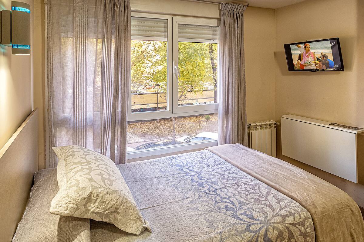 A hotel room with a bed, a window with curtains, a radiator, and a television on the wall.
