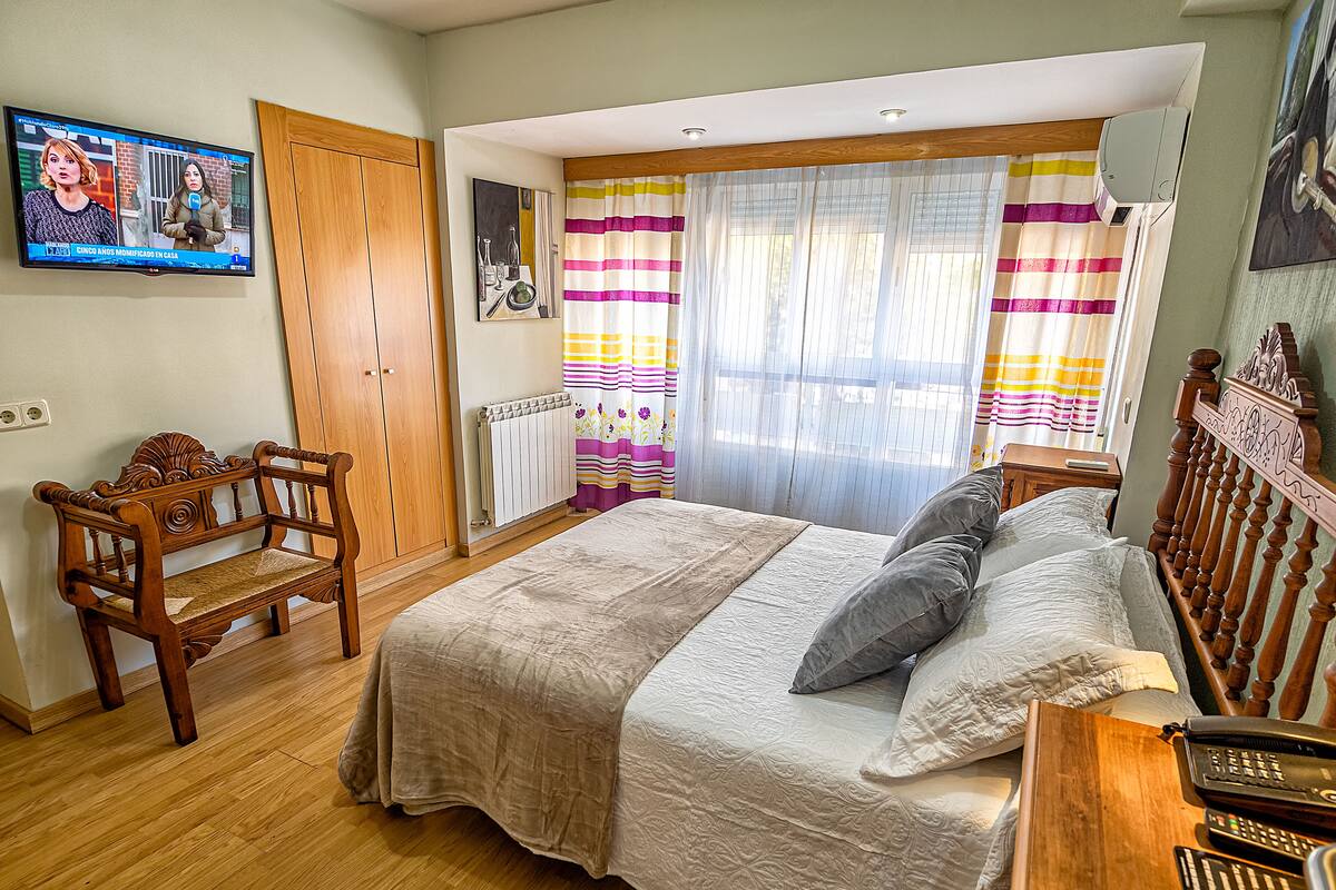 A bedroom with a wooden bed, a wooden chair, a TV on the wall, and a window with curtains.