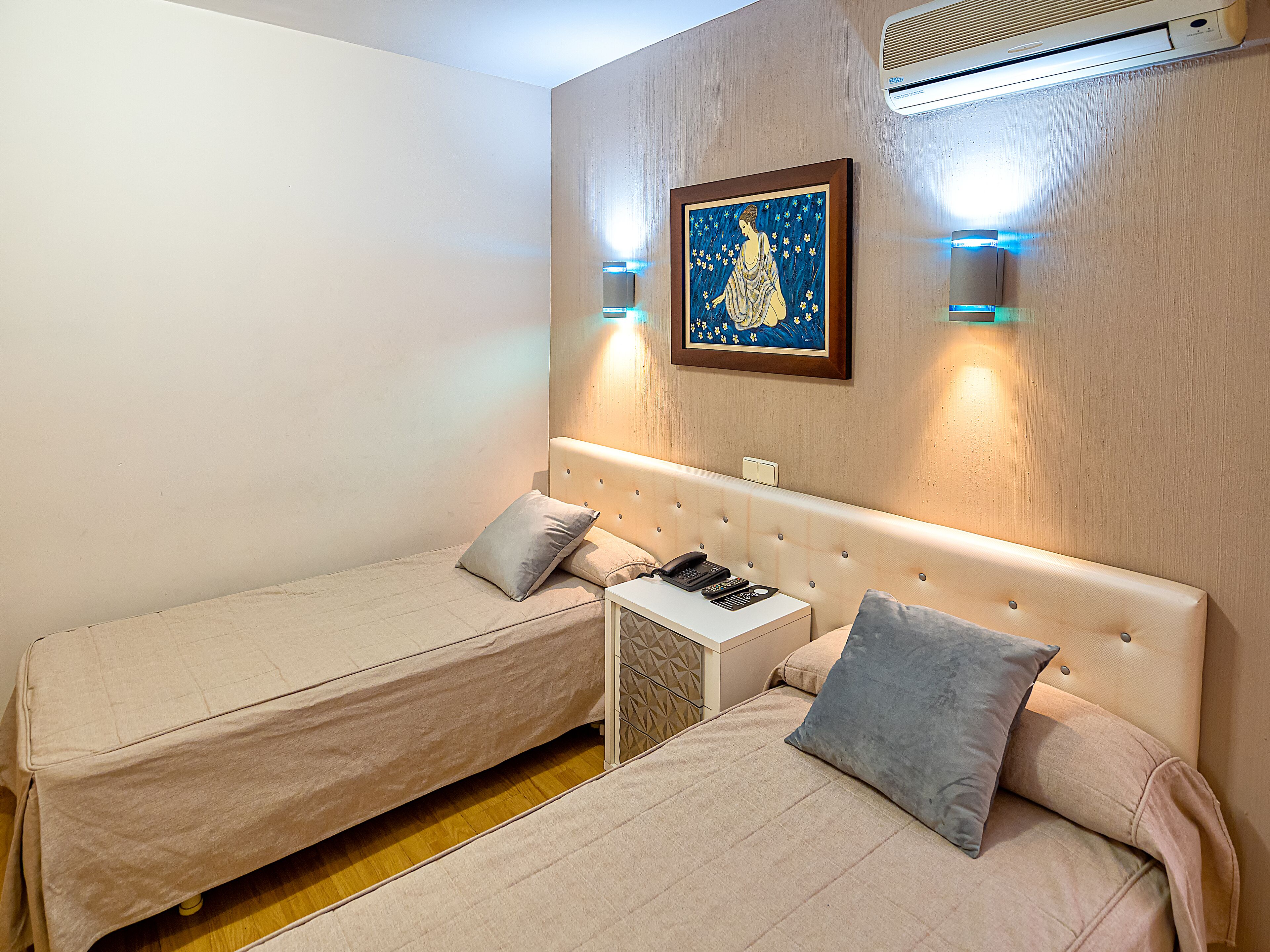 Habitación de hotel con dos camas individuales, una mesita de noche con teléfono y controles remotos, aire acondicionado de pared y una obra de arte enmarcada sobre las camas.