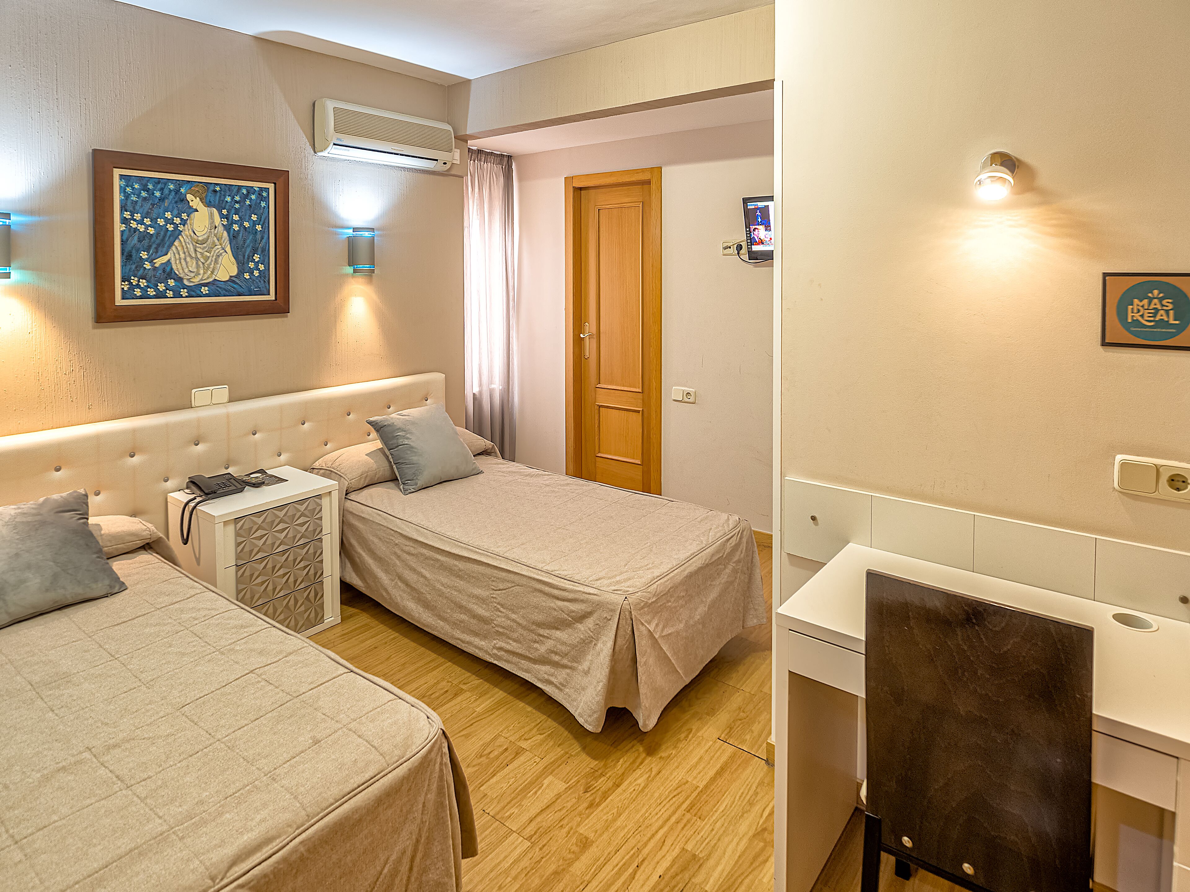 Een hotelkamer met twee bedden, een bureau, een stoel, een airconditioner, een schilderij en een deur.
