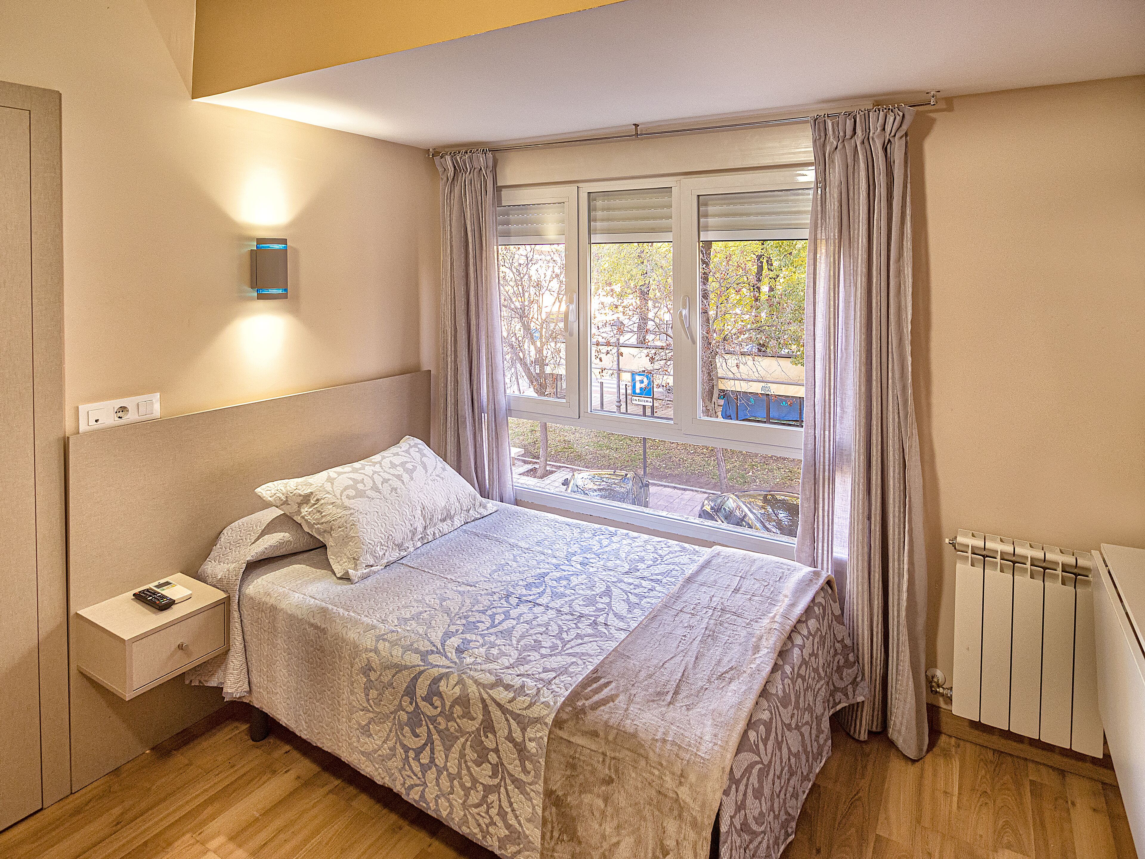 A bedroom with a bed, a nightstand, a window with curtains, and a radiator.