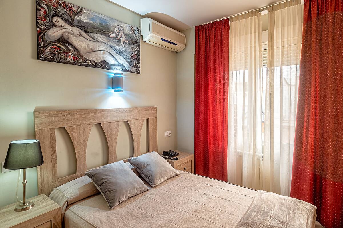 A bedroom with a wooden headboard, a bed with two pillows, a bedside table with a lamp, and a painting on the wall.