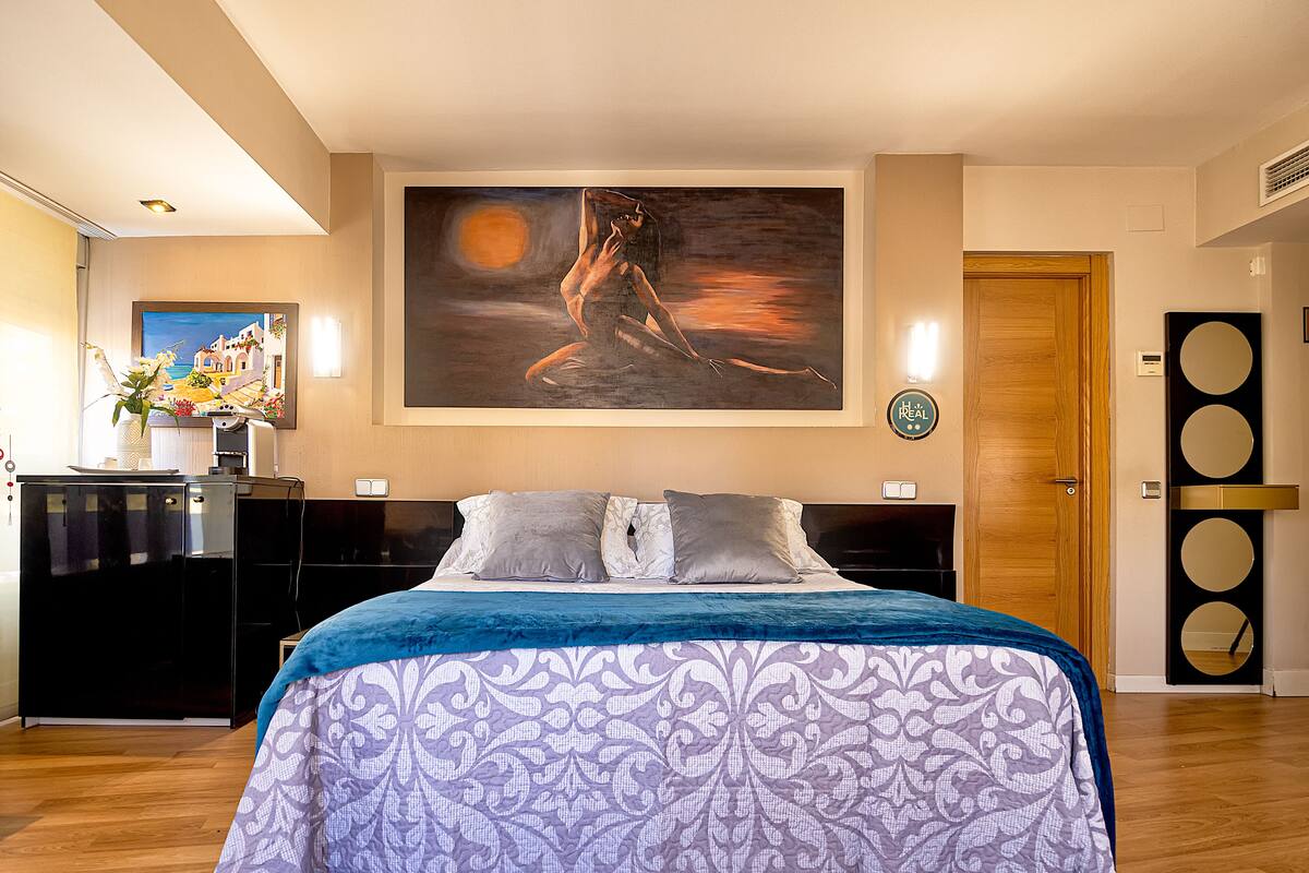 A bedroom with a large bed, a painting on the wall, a wooden door, and a small table with a vase of flowers.