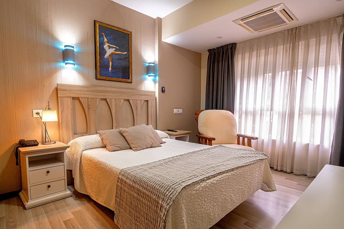 A hotel room with a wooden headboard, a large bed with a textured blanket, a chair, a nightstand with a lamp, and a painting on the wall.