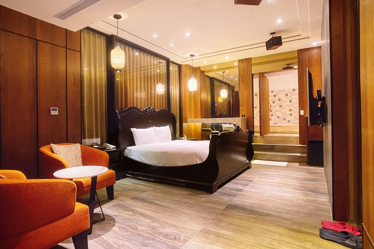 A hotel room with a large bed, a sitting area with a chair and a small table, a mirror, and a bathroom visible through an open door.