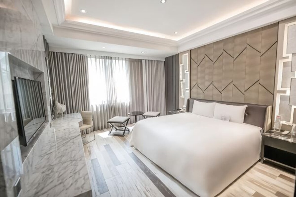 A modern hotel room with a large bed, a TV, a seating area, and a view of the outdoors.