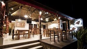 A restaurant with wooden tables and chairs, a stone wall, and a thatched roof. - Villa Puspa (Tabanan)