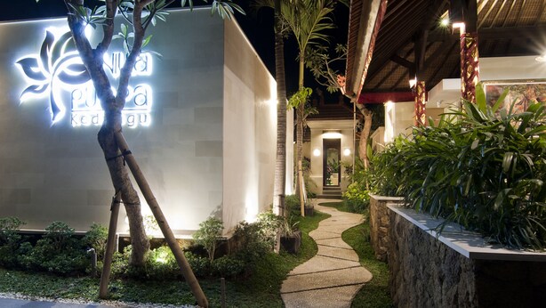 A nighttime view of a building with a pathway leading to the entrance, surrounded by greenery and a stone wall. - Villa Puspa (Tabanan)
