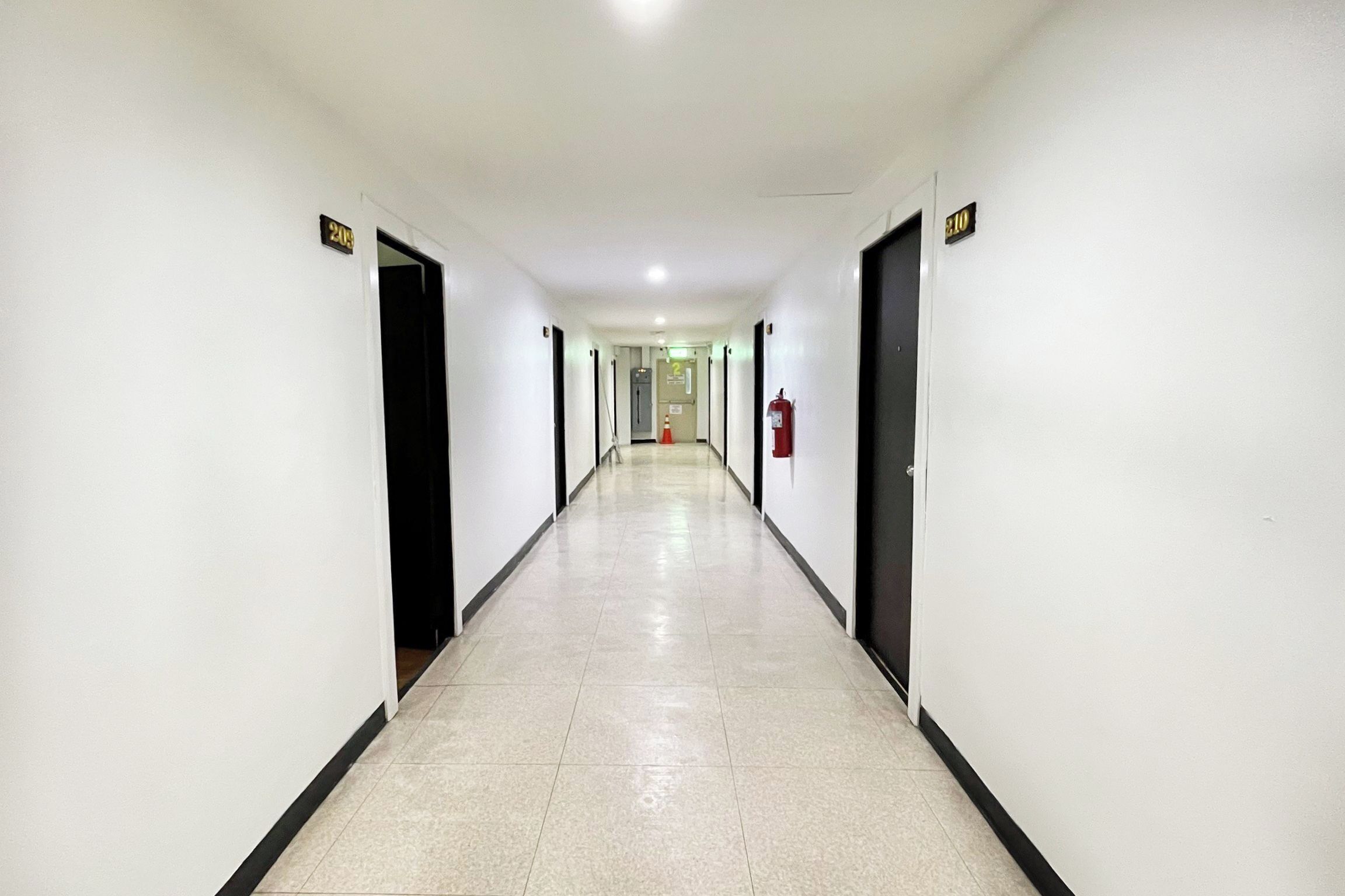 A long, narrow hallway with doors on both sides, a fire extinguisher on the right wall, and a sign indicating room numbers on the left wall.