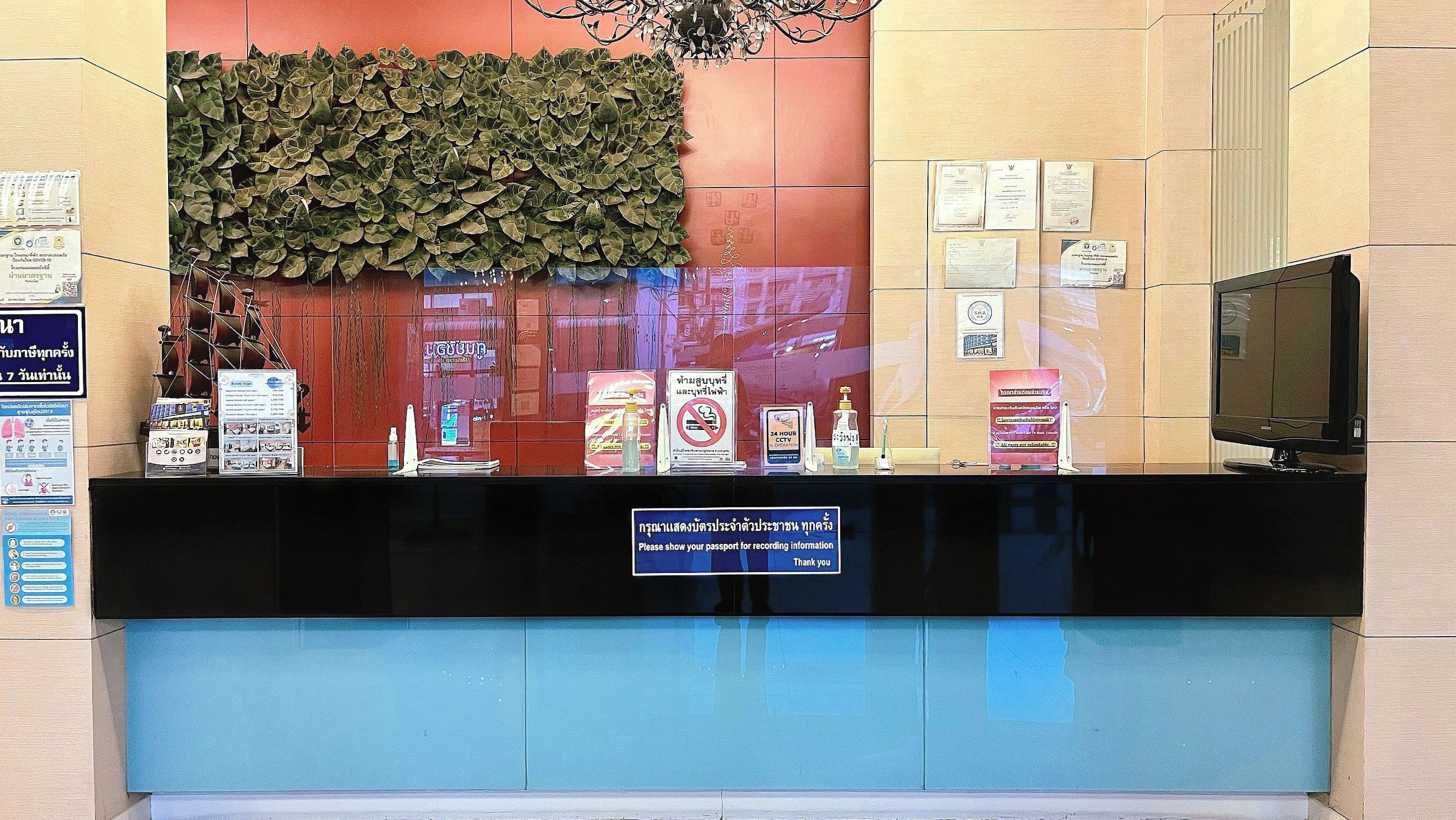 A reception desk with a sign requesting passport display, surrounded by informational posters and a decorative plant wall.