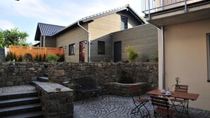 A courtyard with a stone wall, a wooden bench, a table with chairs, and a building with a sloped roof. - Vulkanhotel Balance Selfness (Steffeln)