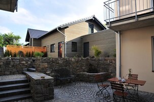 Un patio con un muro de piedra, un banco de madera, una mesa con sillas y un edificio con techo inclinado.
