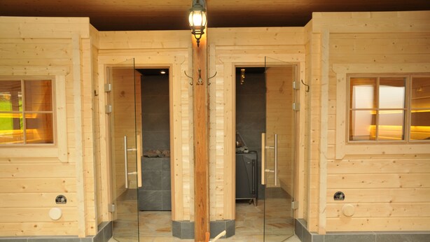 A wooden cabin interior with a central light fixture, glass doors leading to a sauna and a shower room, and a stone-tiled floor. - Vulkanhotel Balance Selfness (Steffeln)