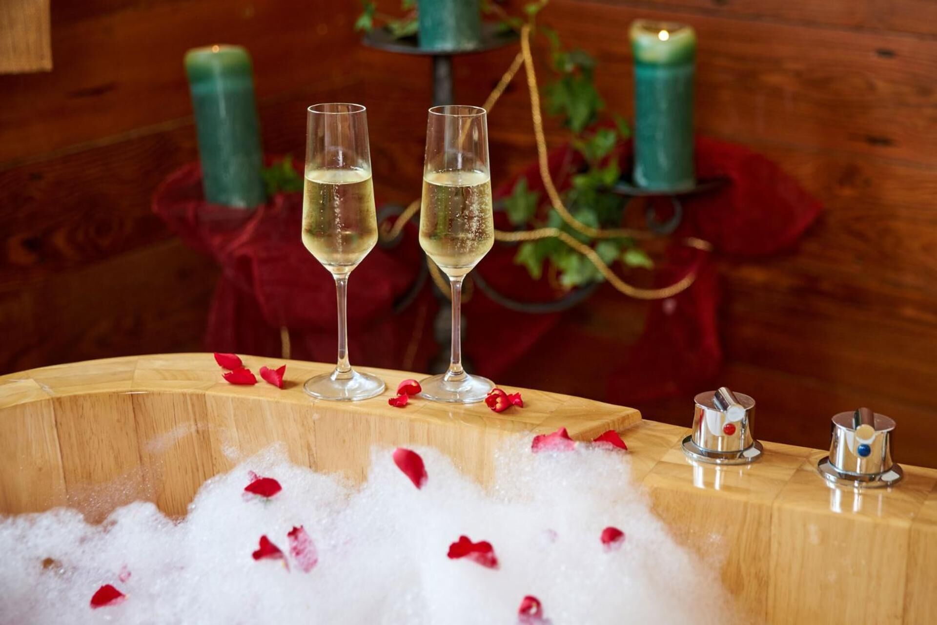 Two glasses of champagne on a wooden table with rose petals, surrounded by candles and a decorative vase.