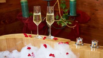 Two glasses of champagne on a wooden table with rose petals, surrounded by candles and a decorative vase.