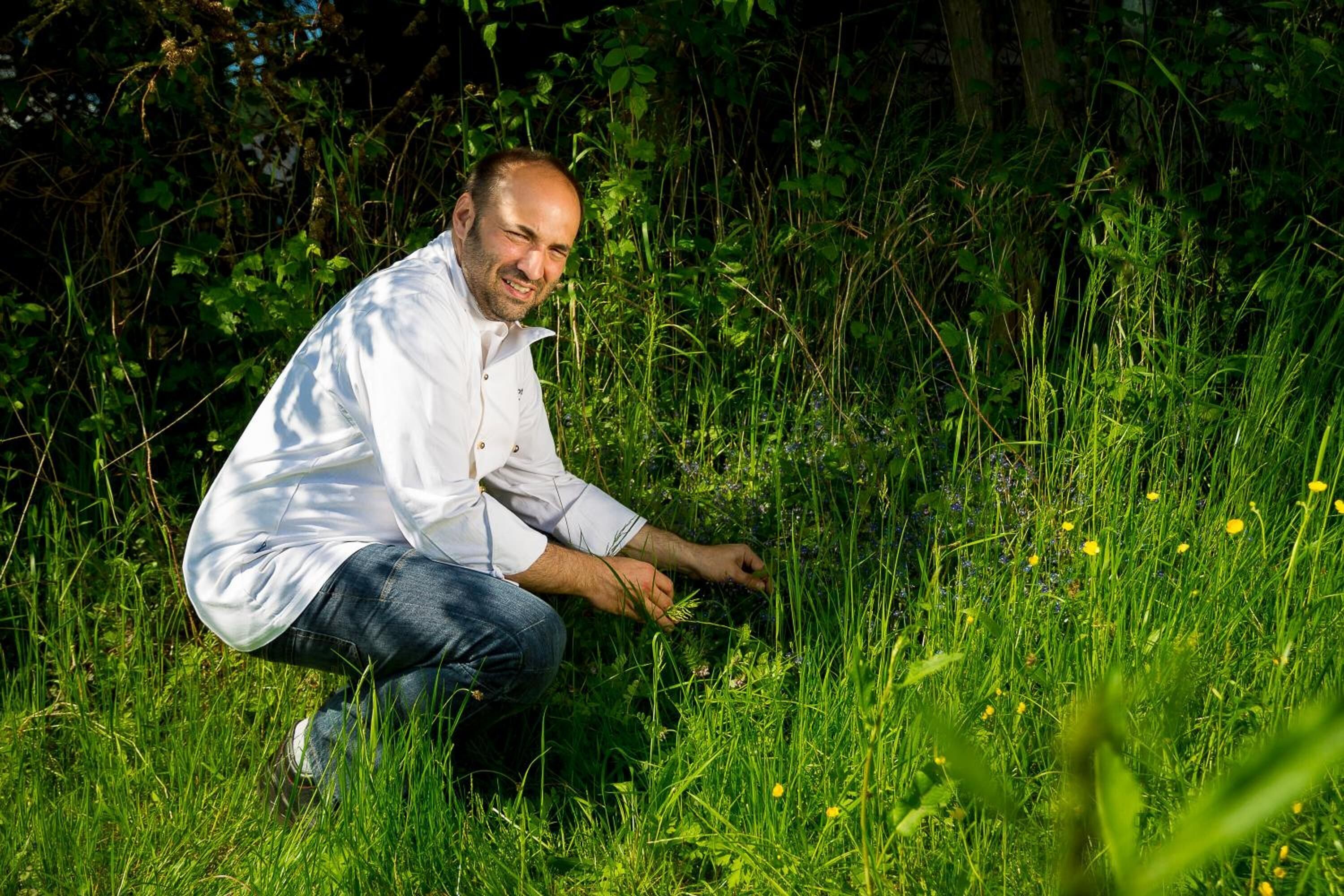 Una persona agachada en una zona cubierta de hierba, con vegetación densa y árboles al fondo.