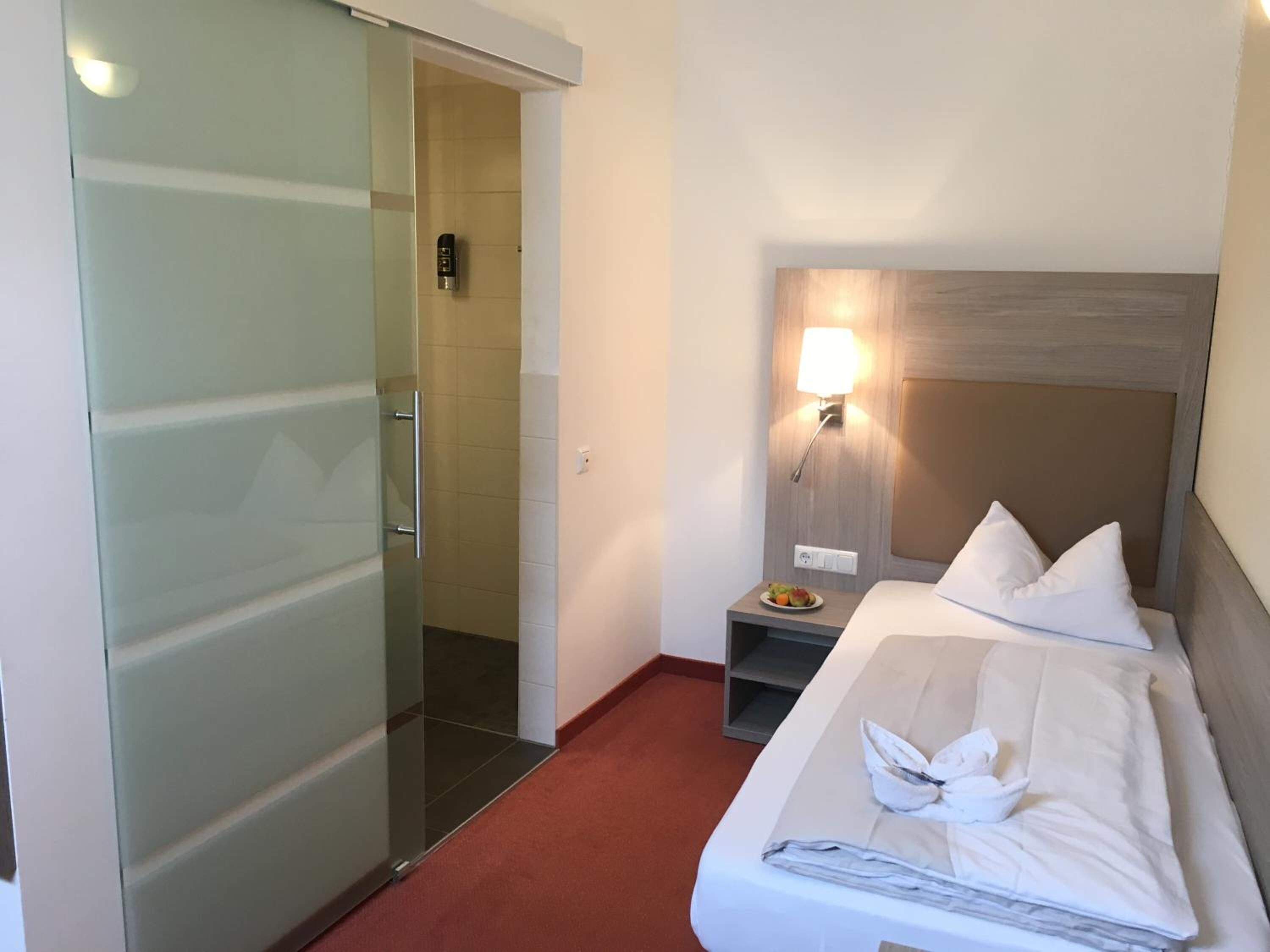 A hotel room with a bed, a nightstand, a wall-mounted lamp, and a sliding door leading to a shower area.