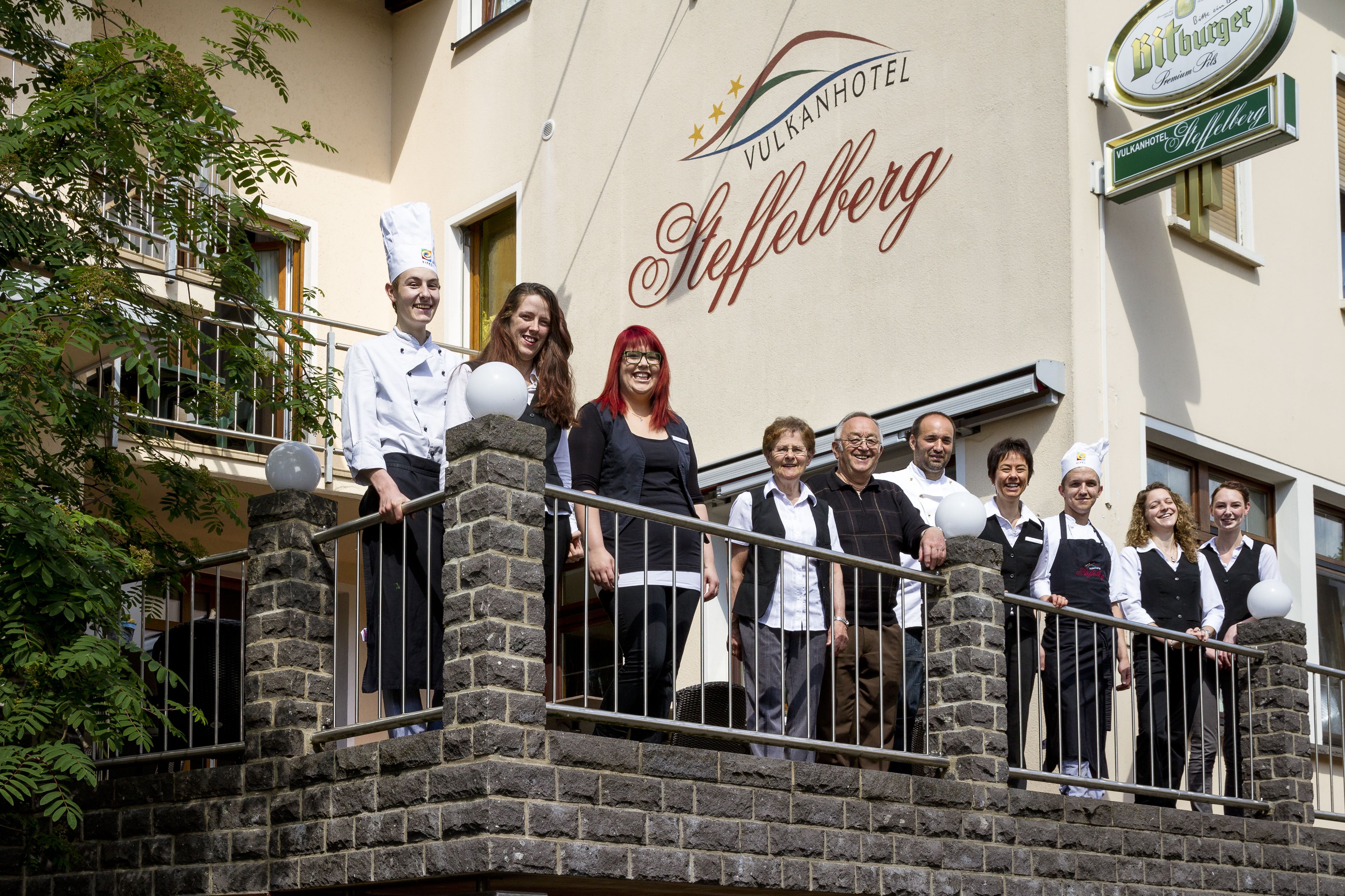 Eine Gruppe von Menschen steht auf dem Balkon eines Hotels, auf dem ein Schild mit der Aufschrift "Vulkanhotel Steffelberg" zu sehen ist.
