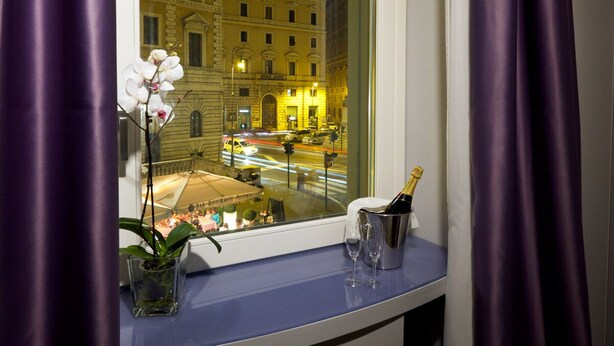 A window with a view of a city street at night, a vase with white flowers, a bottle of champagne, and a glass. - Navona Rooms (Rome)
