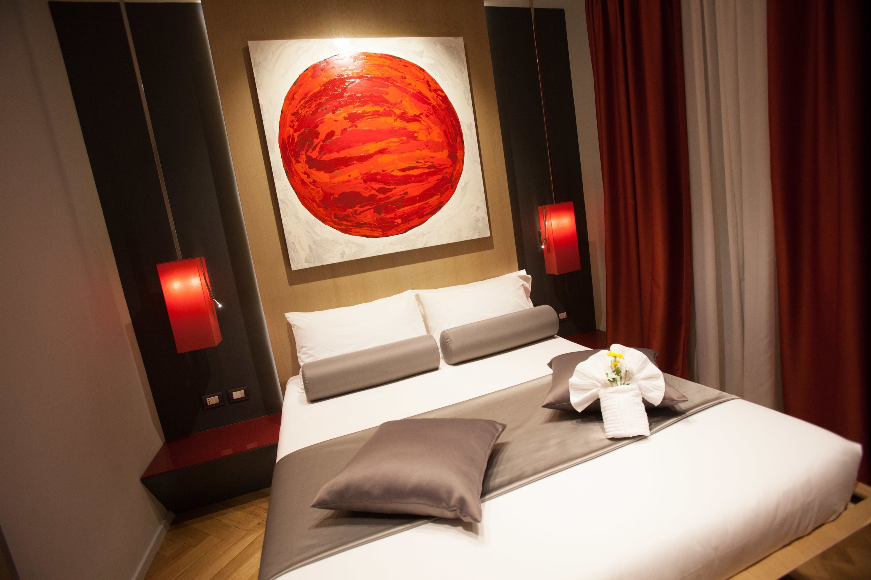 Une chambre d’hôtel avec un grand tableau représentant une planète, un lit avec une literie grise et un éclairage d’ambiance rouge.