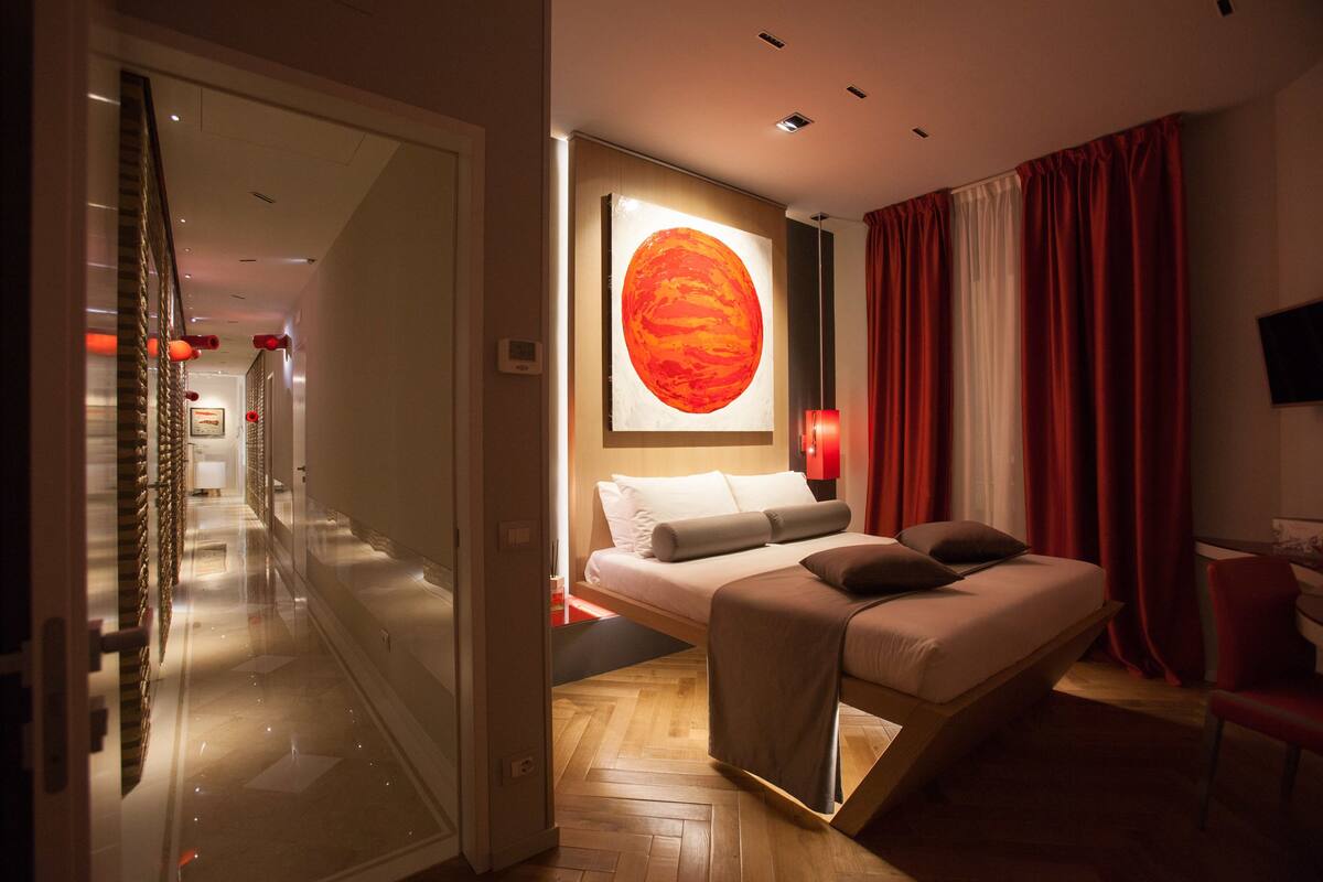 A modern hotel room with a bed, a wall-mounted TV, and a large window with red curtains.