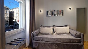 A modern living room with a grey sofa, a small table, and a window with a view of buildings. - Navona Rooms (Rome)