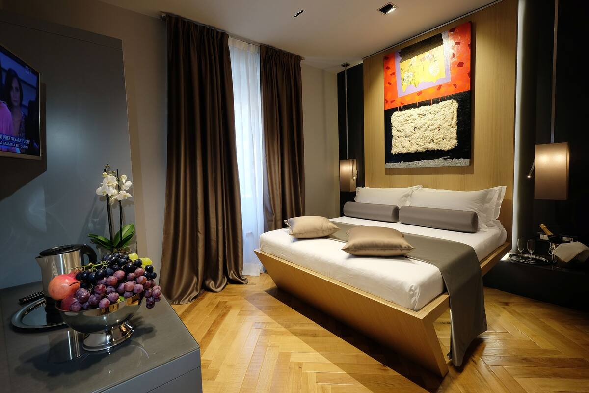 A modern hotel room with a large bed, a flat-screen TV, a fruit bowl, and a decorative wall piece.