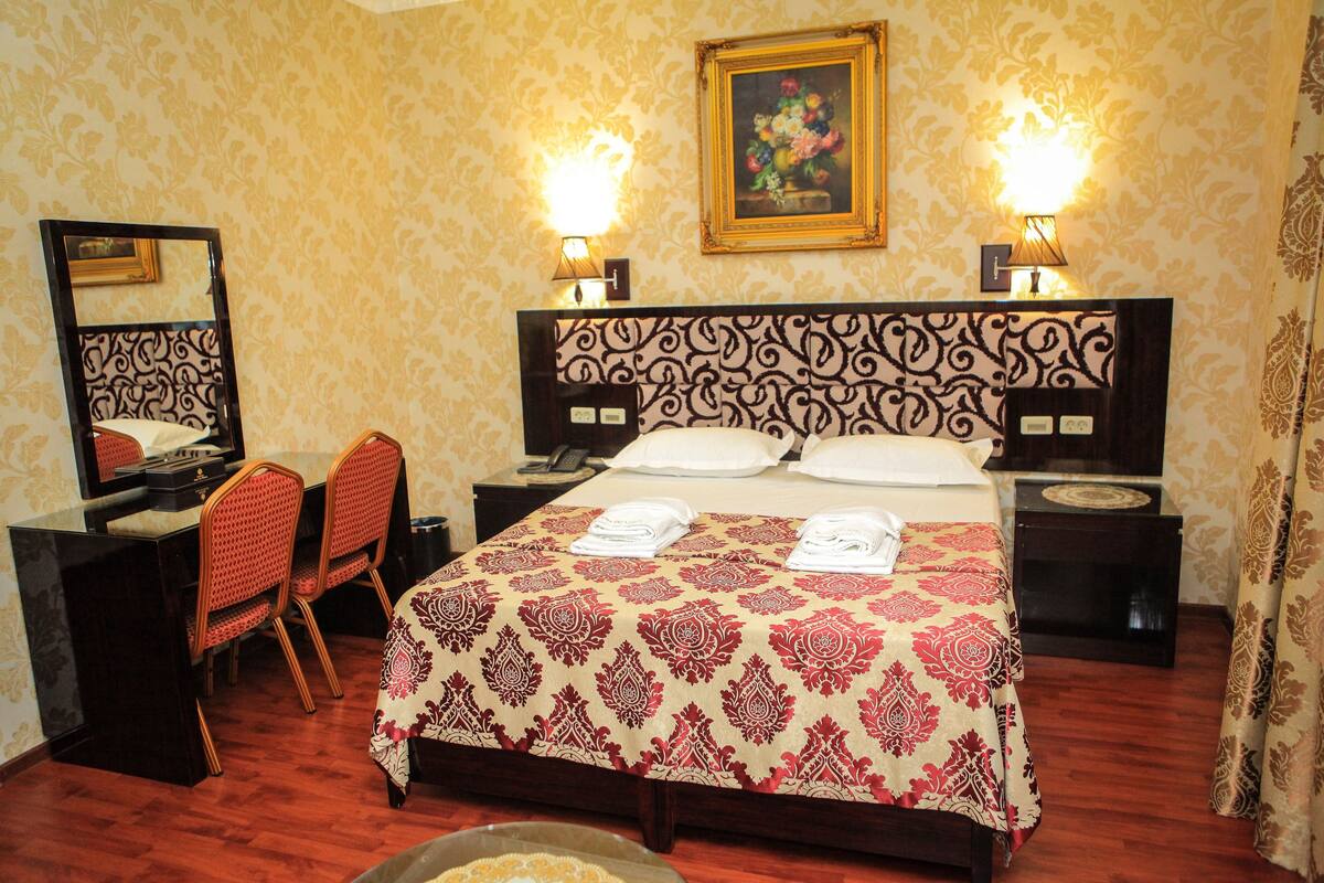 A hotel room with a large bed, two bedside tables, a desk with a chair, a mirror, and two wall-mounted lamps.