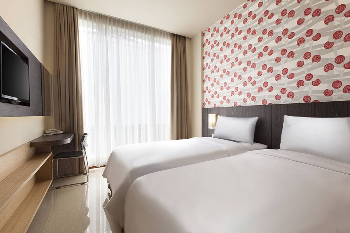 Ein Hotelzimmer mit zwei Betten, einem an der Wand montierten Fernseher, einem Schreibtisch mit Telefon und einem Fenster mit Vorhängen.