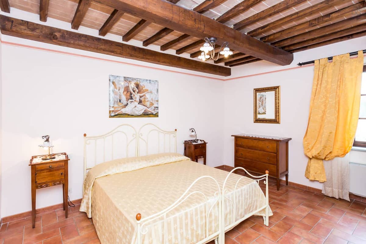Een slaapkamer met een bed, nachtkastje, dressoir, schilderij aan de muur en een raam met gordijnen.