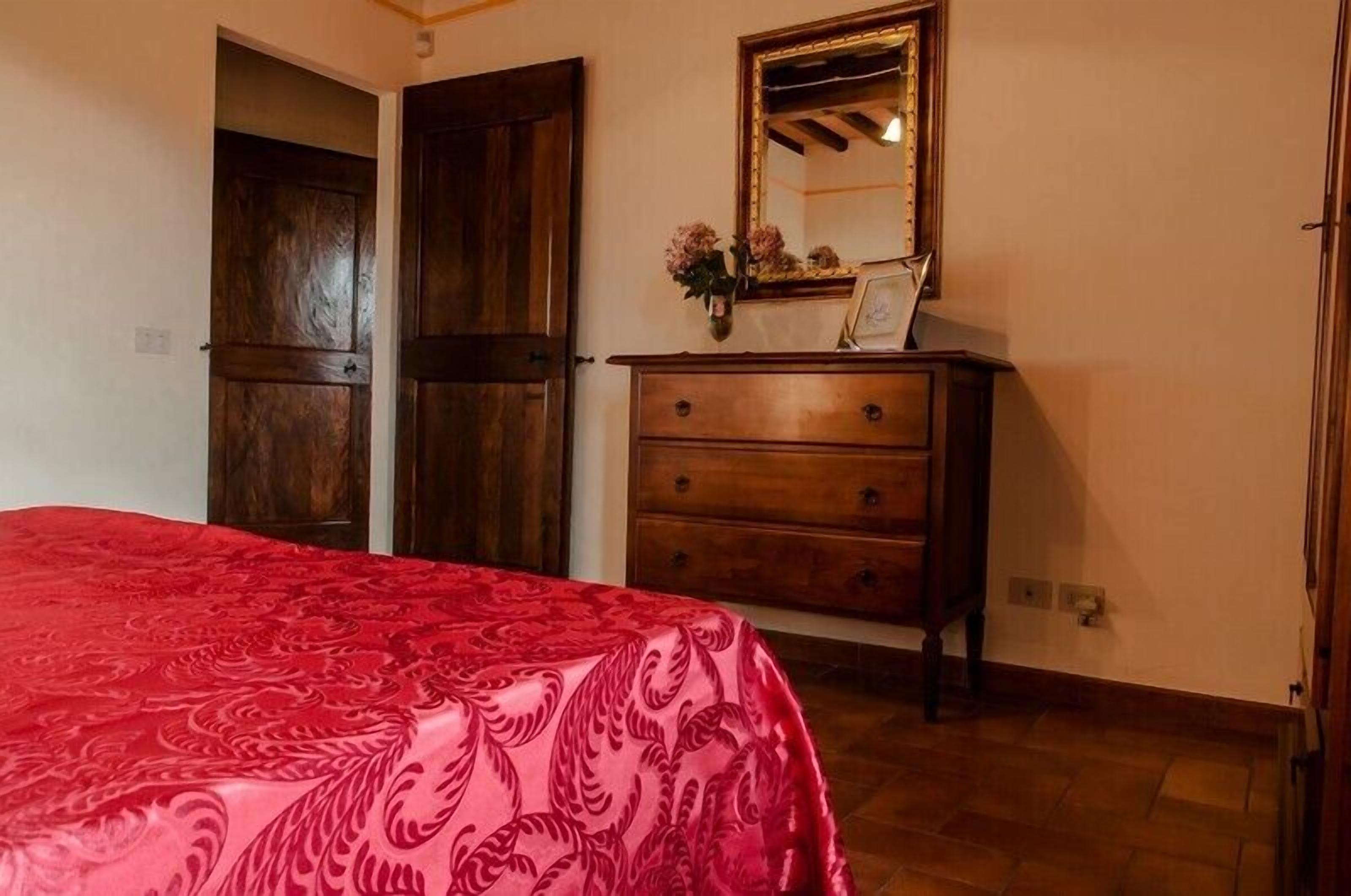 A bedroom with a red patterned bedspread, a wooden dresser, a mirror, and a door.