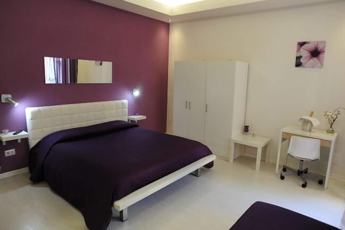 Un dormitorio con cama, mesita de noche, armario, escritorio, silla y una pequeña mesa con un florero.