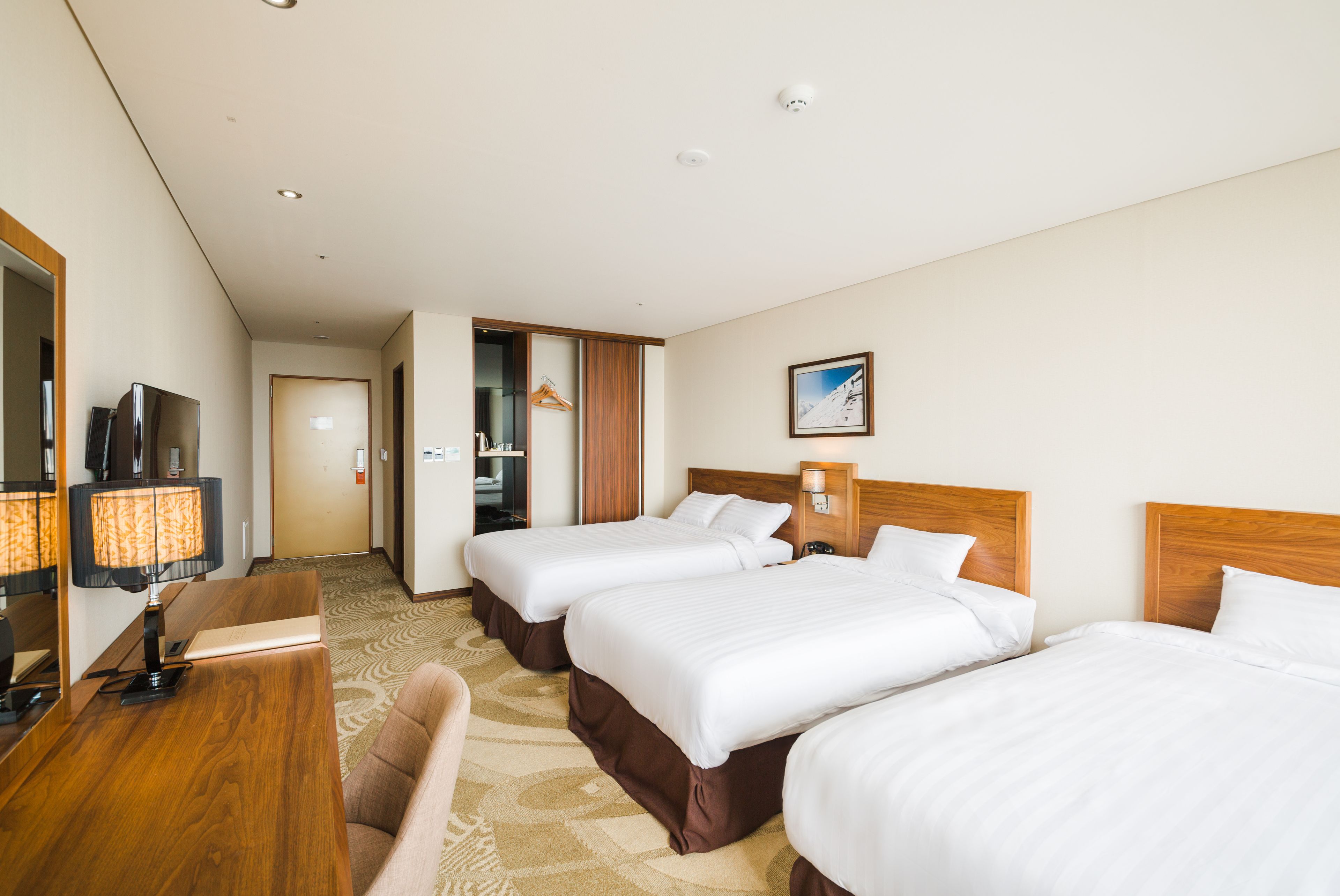 A hotel room with three beds, a desk with a lamp, a chair, a coat rack, and a framed picture on the wall.