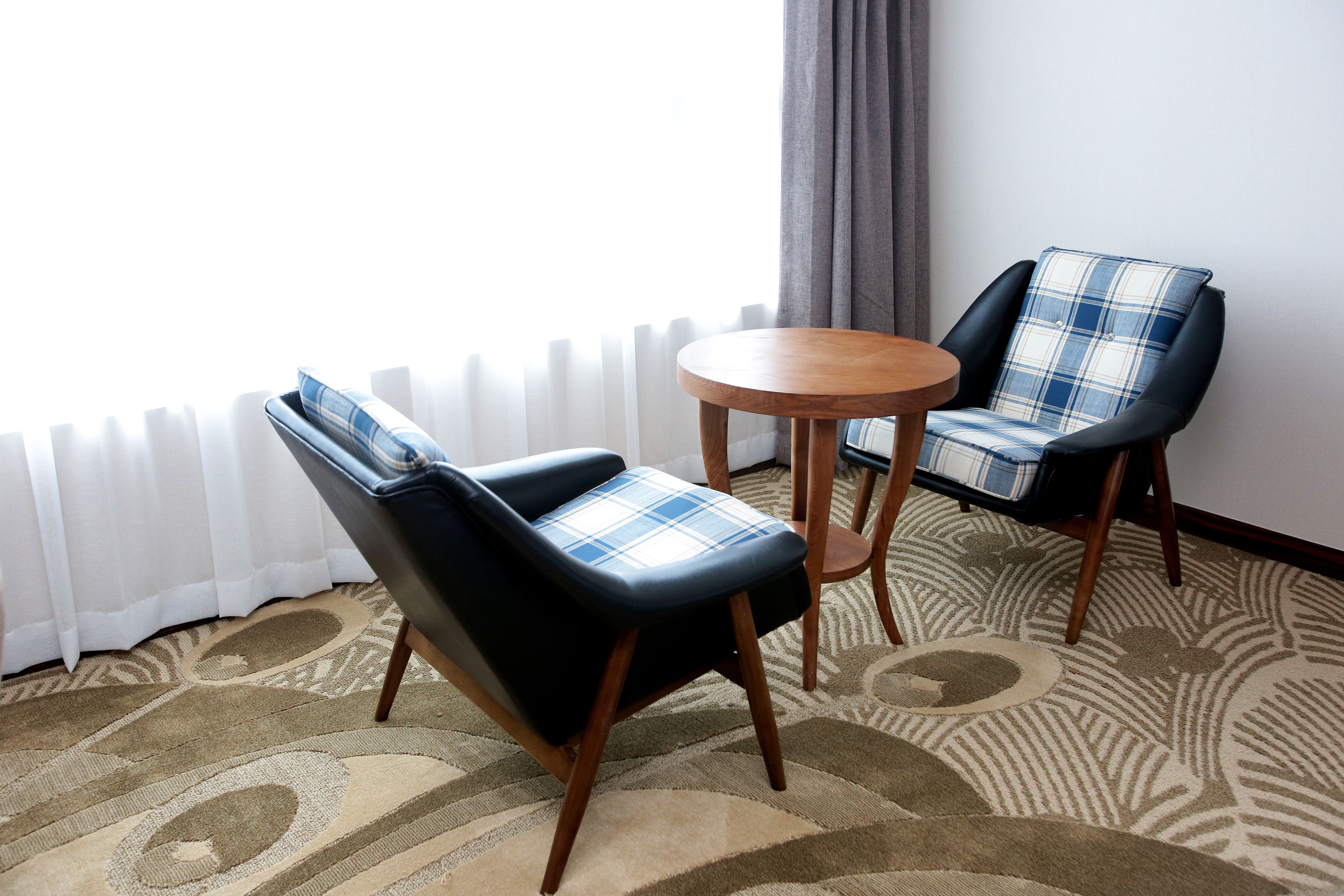 Twee stoelen met geruite kussens, een klein rond tafeltje en een kamer met een radiator, gordijnen en een vloerkleed met dessin.