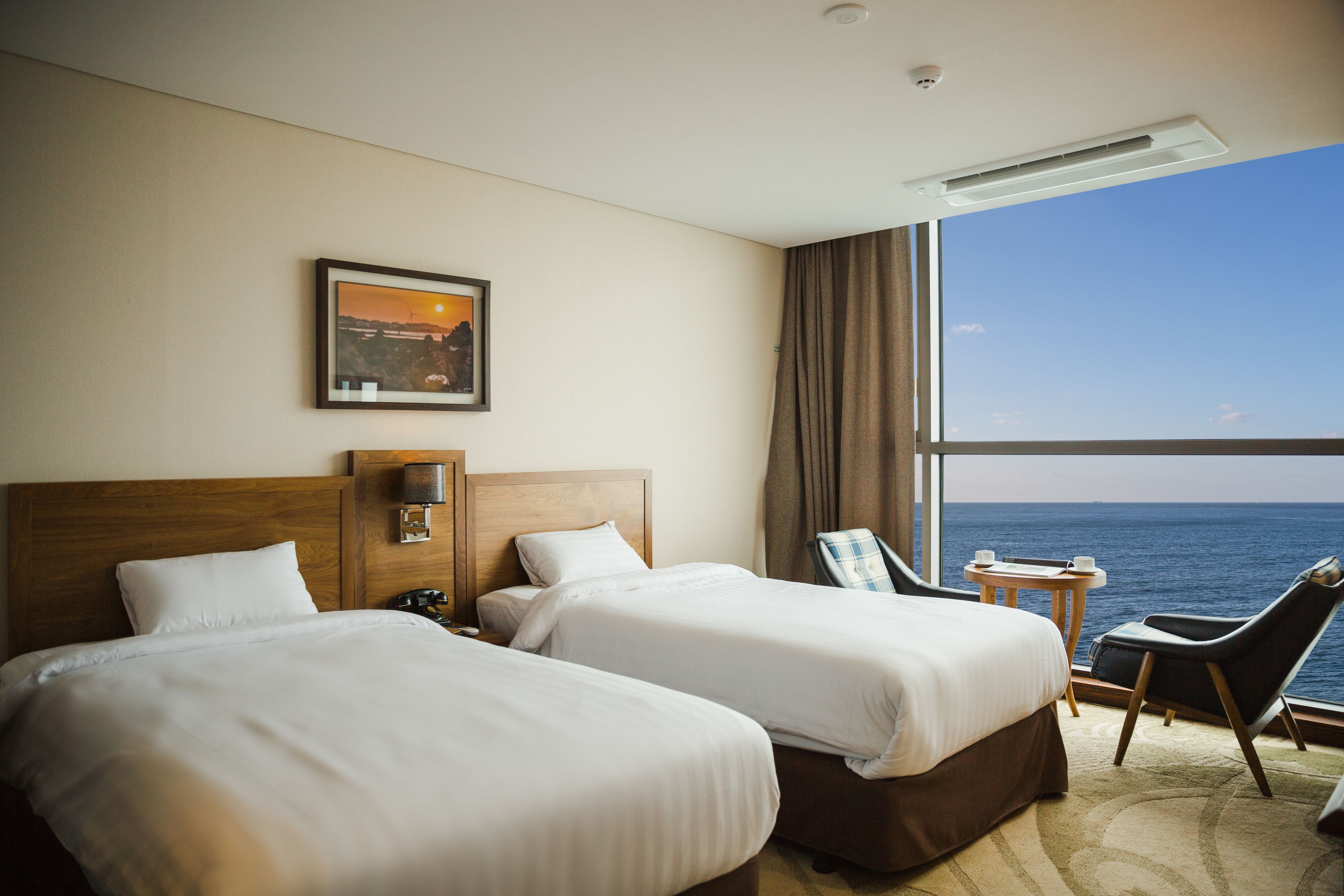 Een hotelkamer met twee bedden, een kleine tafel, een stoel en uitzicht op zee.