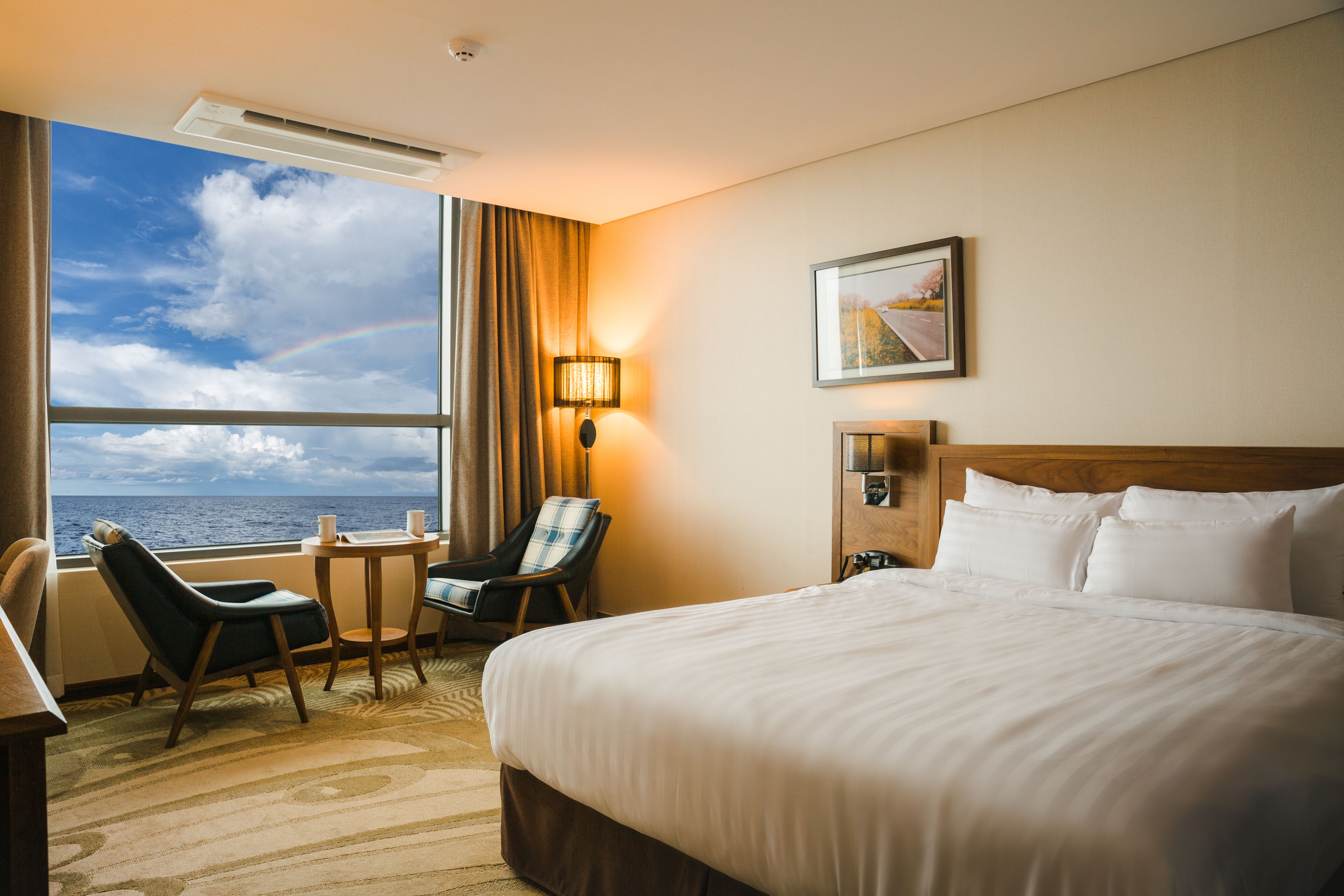 A hotel room with a large bed, a chair, a small table, a lamp, and a window with a view of the sea.
