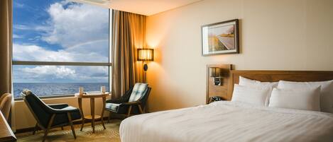 A hotel room with a large bed, a chair, a small table, a lamp, and a window with a view of the sea.