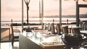 A dining area with a view of a body of water and city skyline, featuring a long table with chairs and various dishes. - Tamna Stay Hotel Jeju (Jeju City)