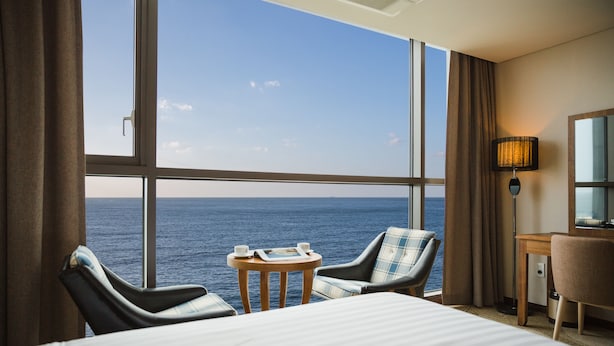 A hotel room with a large window overlooking the sea, a bed, two chairs, a small table with a magazine and a cup, and a desk with a lamp. - Tamna Stay Hotel Jeju (Jeju City)