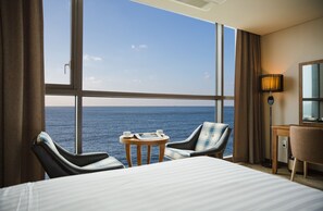 Une chambre d’hôtel avec une grande fenêtre donnant sur la mer, un lit, deux chaises, une petite table avec un magazine et une tasse, et un bureau avec une lampe.