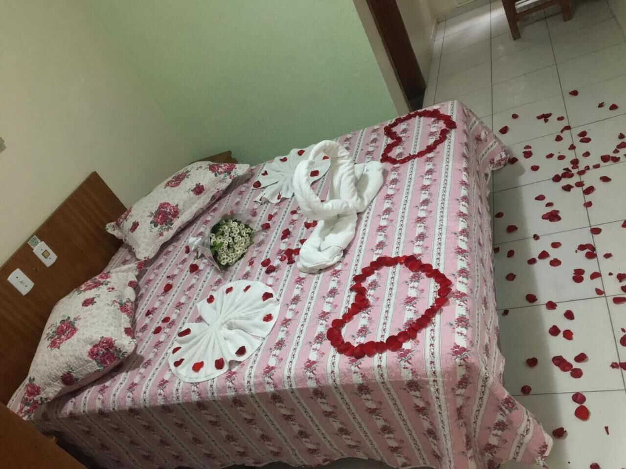 Un lit recouvert d’une couvre-lit à motifs floraux, un arrangement en forme de cœur composé de pétales de roses et une serviette blanche.