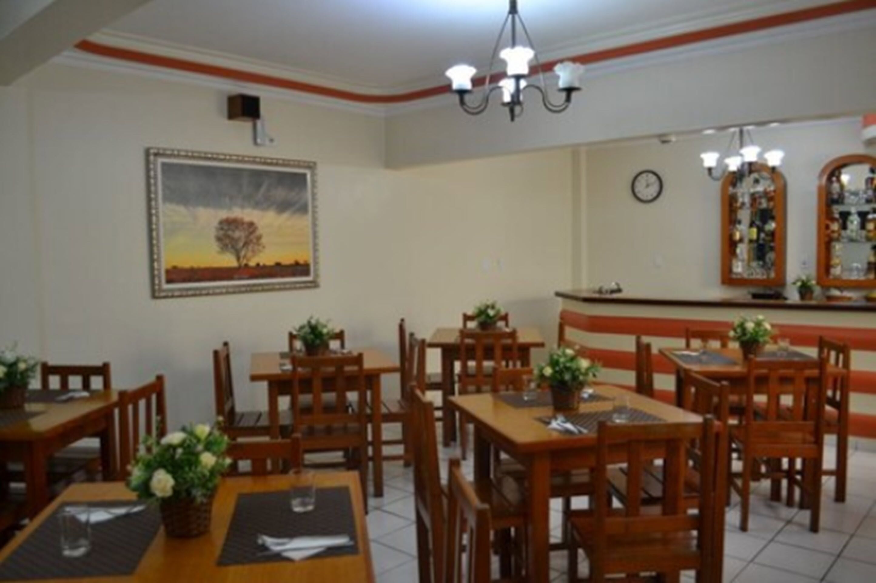 Un restaurante con mesas y sillas de madera, un cuadro en la pared y una zona de bar con botellas.
