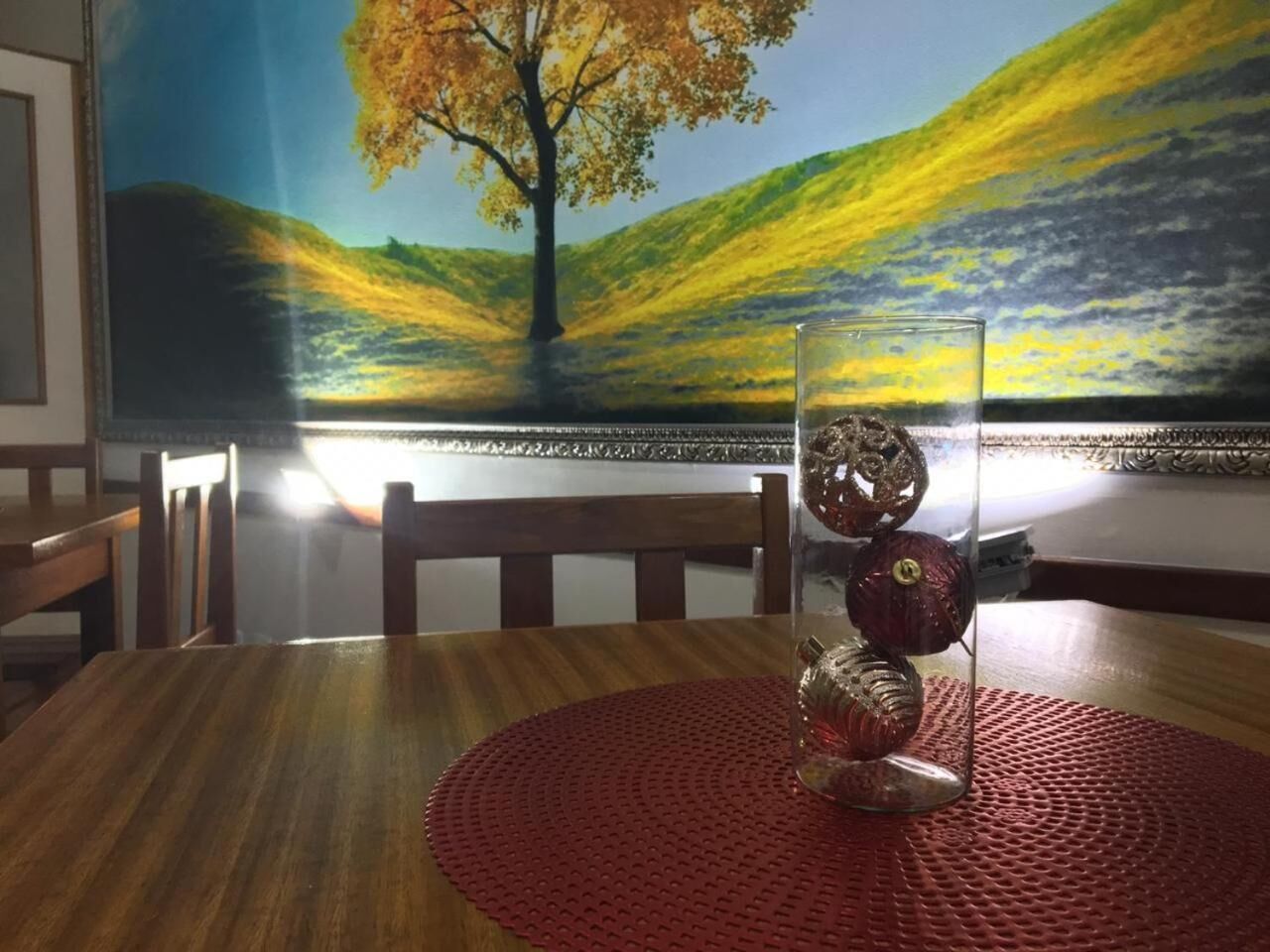 A dining table with a clear glass vase containing decorative items, a red placemat, and a painting of a tree on the wall behind.