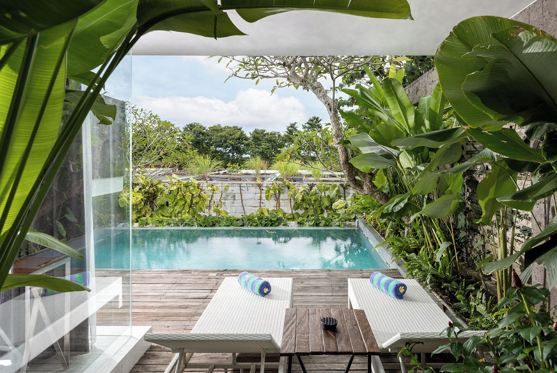 Uma área de piscina com duas espreguiçadeiras e uma mesa de madeira, cercada por vegetação exuberante e vista para um jardim.