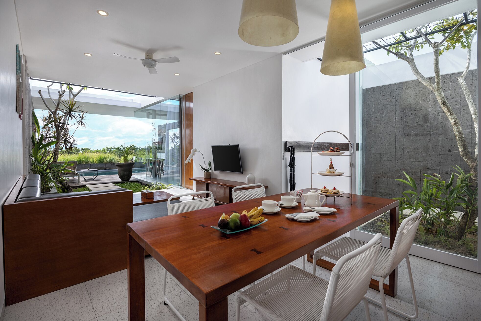A modern dining area with a wooden table, white chairs, and a fruit bowl. There is a TV on the wall and a plant in the corner.
