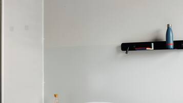 A neatly made bed with white linens, a bedside table with a cup and a bottle, and a wall-mounted shelf with a blue water bottle and a book.