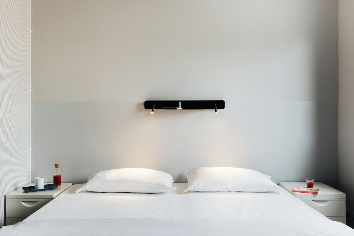 Habitació d'hotel minimalista amb un llit, dos coixins blancs, una taula de nit amb un got d'aigua, una ampolla i un safata.