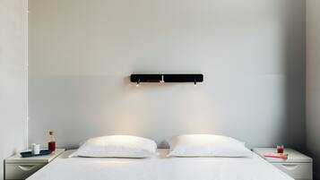 Camera d'albergo dal design minimalista con un letto, due cuscini bianchi, un comodino con un bicchiere d'acqua, una bottiglia e un vassoio.
