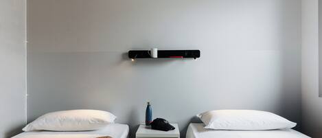 Un dormitorio con dos camas individuales, una mesita de noche con una lámpara, un estante pequeño con una taza y una botella de agua.